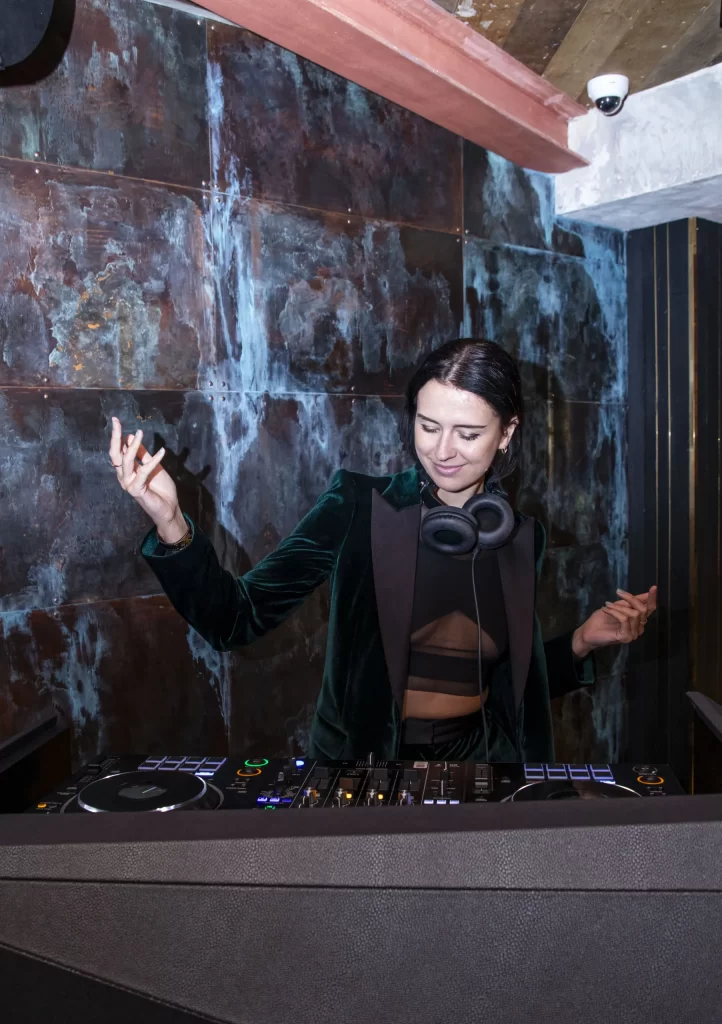 A woman is DJing in front of a DJ booth at a restaurant near Harrods.